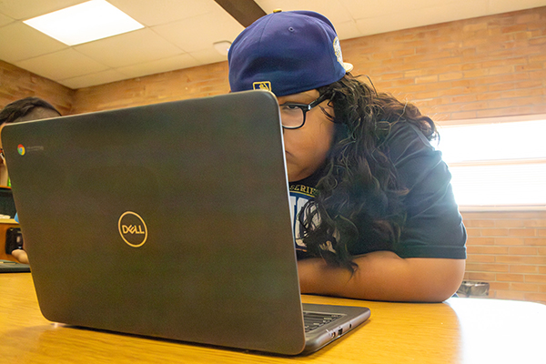 A boy wearing glasses and a backwards hat works on his laptop